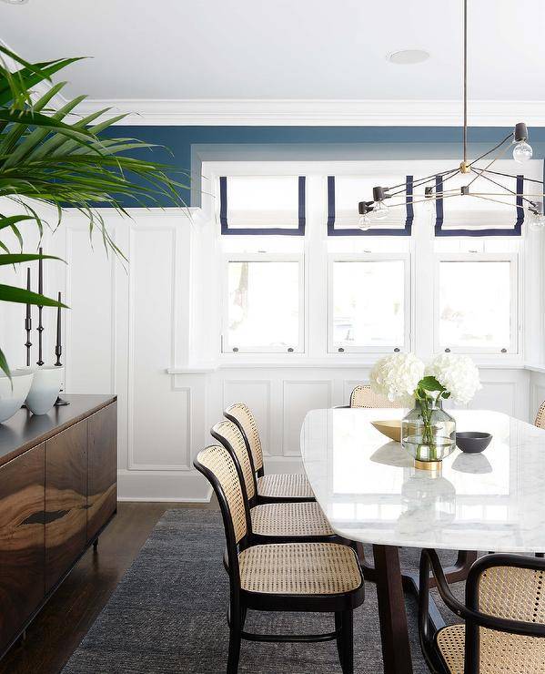 Blue grosgrain roman shades cover a row of a window framed by a board and batten wall finished with a blue upper wall in a charming cottage dining room. Cane chairs are placed on a charcoal gray rug at a marble top dining table.