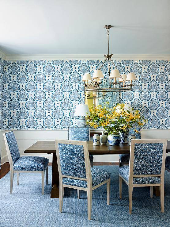 Galbraith & Paul Lotus wallpaper lined with white wainscoting accents a beautiful blue dining space featuring a brown trestle table surrounded by gray and blue chairs placed on a blue rug and lit by a candelabra chandelier.