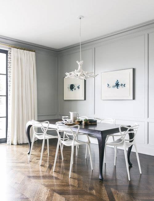 Beautifully designed contemporary dining room is lit by a white faux antlers chandelier hung over a black French dining table surrounded by Kartell Masters Chairs placed on a wood herringbone floor. Blue art hangs on a gray wall accented with gray paneling and gray crown molding, while white French pleated curtains hang in front of black glass doors.