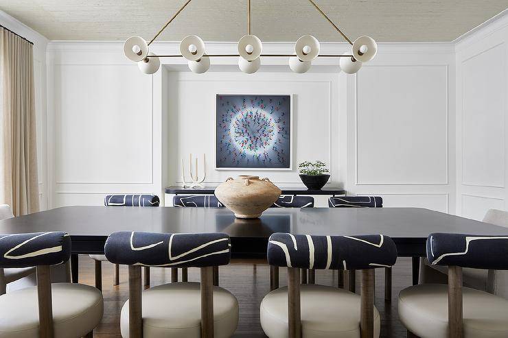 Cream and black graphic chairs sit at a rectangular black dining table lit by a white and gold linear chandelier. Wainscoting frames a small alcove featuring a black and white curved buffet cabinet placed beneath a square frame art piece.