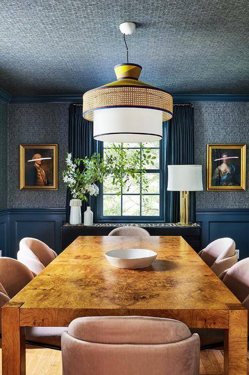 Hung from a blue wallpapered ceiling, a vintage cane lantern lights a burl wood dining table matched with pink velvet chairs. Gold framed vintage art hangs on either side of a window from blue wallpapered walls lined with blue wainscoting.