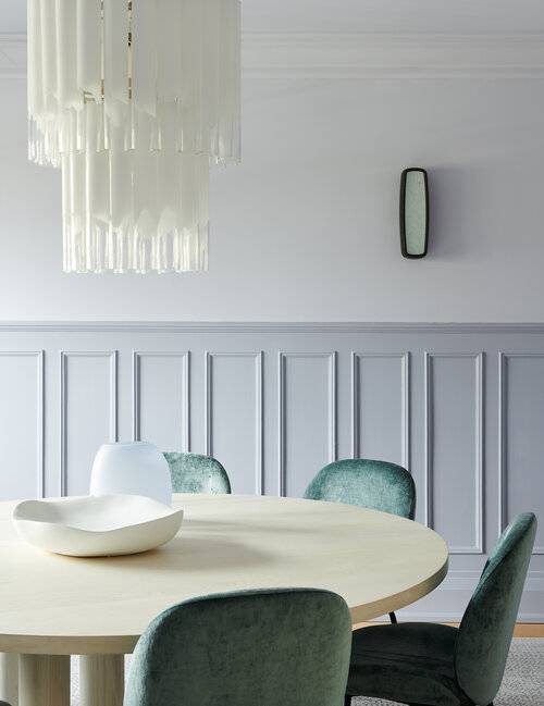 Half wainscot dining room walls are positioned behind a round cream dining table matched with green velvet dining chairs and lit by a white cascading chandelier.