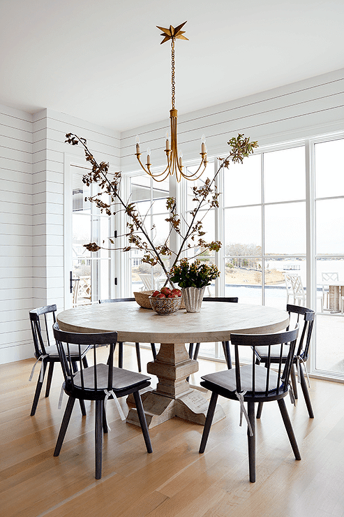 A brass chandelier hangs over a round, brown dining table paired iwth black spindle dining chairs. The dining space is positioned on a stained wood floor in front of windows framed by white shiplap.