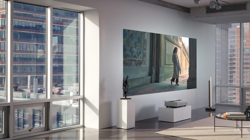 Contemporary hi-rise loft living room with Leica Cine 1 projector set on a low white pedestal console projecting large 100-inch movie with woman in long sweater walking around somewhere in Europe,