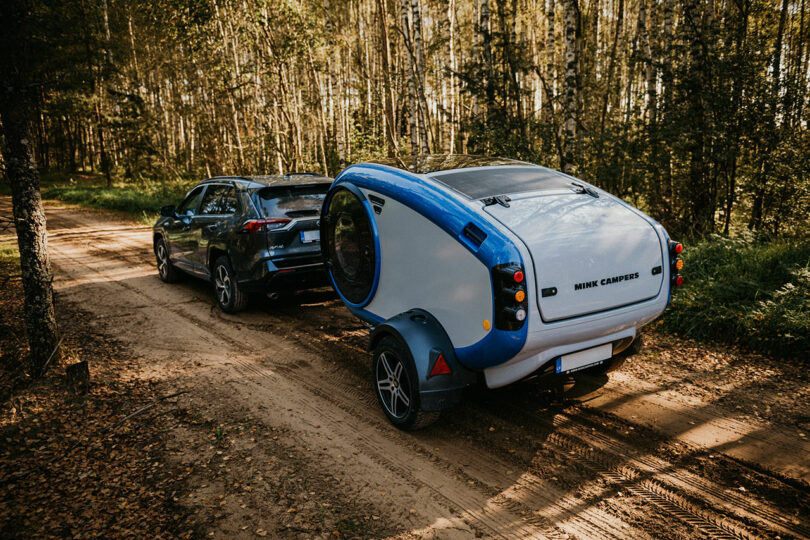 back angled view of modern gray and blue teardrop camper pulled by suv in woods
