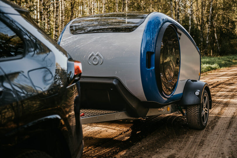 angled view of modern gray and blue teardrop camper pulled by suv in woods