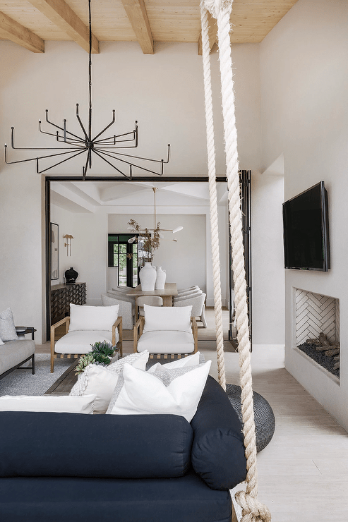 Hung from a wood plank patio ceiling, a large oil rubbed bronze chandelier illumates a black rope swing sofa hung facing a gray teak coffee table and beige teak accent chairs. A gray industrial fireplace is finished with a flat panel TV.