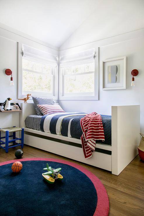 A white twin bed with trundle designed in a nautical theme dressed in red, white and blue striped bedding. Corner bedroom windows dressed with white roman shades are flanked by red industrial sconces complimenting a round red and navy blue rug layered with wood floors.