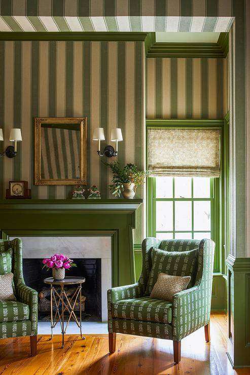 This gorgeous green formal living space features a green fireplace mantel framing a marble surround and fixed against a wall clad in green vertical stripe wallpaper. A gold French mirror hangs above the fireplace and between oil-rubbed bronze 2-light sconces. A brass directoire accent table is flanked by green wingback chairs.