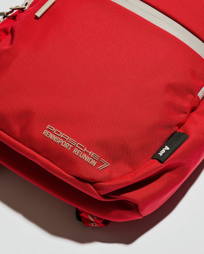 Detail of red Aer x Porsche Rennsport Reunion 7 backpack, showing the "Porsche Rennsport Reunion 7" branding logo in detail and Aer tag.