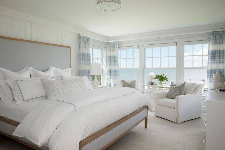 Brown and blue linen bed with white hotel bedding accented with blue borders in a cottage ocean view master bedroom. White linen skirted accent chairs join a white side table at the bedroom windows boasting a stunning view of a serene ocean and clear skies.