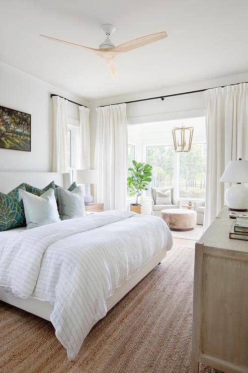 Welcoming master bedroom sitting area features a white curtain hung from an oil rubbed bronze rod in front of a nook boasting a round ottoman placed in front of dove gray wingback chairs lit by a brass lantern hung in front of windows.