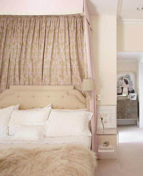 Romantic beige and pink bedroom features a pink and beige pleated valance with a beige and pink canopy accenting a beige tufted headboard positioned behind a bed dressed in a white and beige bedding topped with a beige faux fur throw blanket. Beside the bed, a small acrylic bedside table is illuminated by a nickel wall sconce accented with a beige shade.