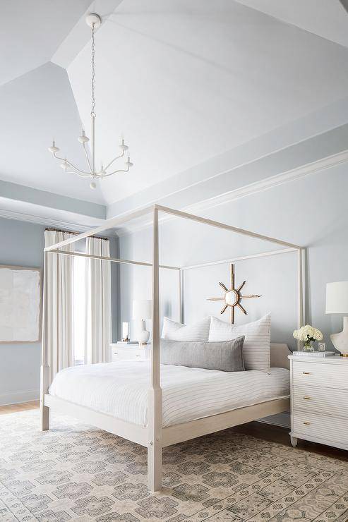 Blue and gray master bedroom design features a light gray wash canopy bed with a gray lumbar pillow and stripe bedding under a gold sunburst mirror, a white French reeded bedside table and a gray vintage rug.