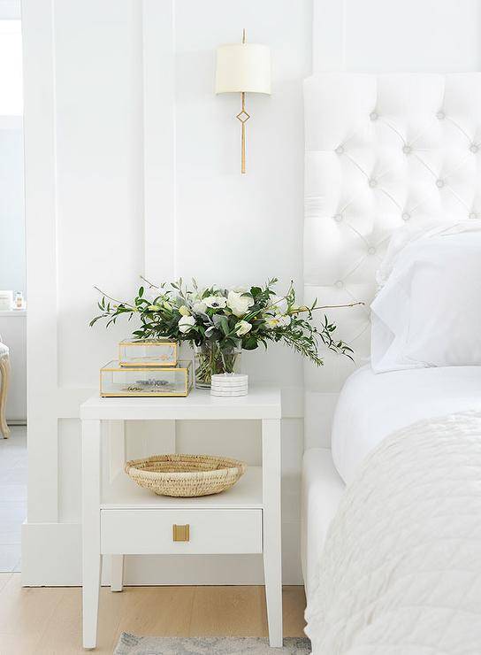 White master bedroom design features a white velvet tufted headboard with a white nightstand and a brass sconce for a warm sophisticated finishing touch.
