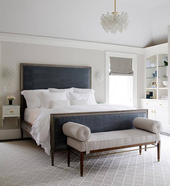 Blue and gray bedroom design with light gray walls framing blue velvet headboard flanked by art deco sconces over white wall-mounted nightstands with brass drawer pulls over gray trellis rug. Gray and blue bedroom features art deco chandelier over blue velvet bed paired with soft white romantic bedding and upholstered gray bench at foot of bed. Master bedroom with wall of white built in cabinets accented with brass hardware and gray woman shade.