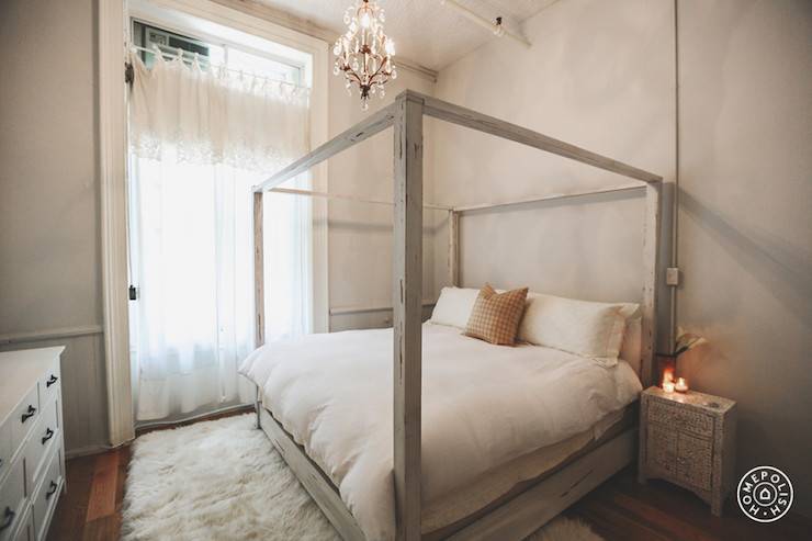 Romantic bedroom features a distressed gray canopy bed dressed in a soft white duvet illuminated by a small crystal chandelier flanked by bone inlay nightstands atop a fluffy white sheepskin rug.