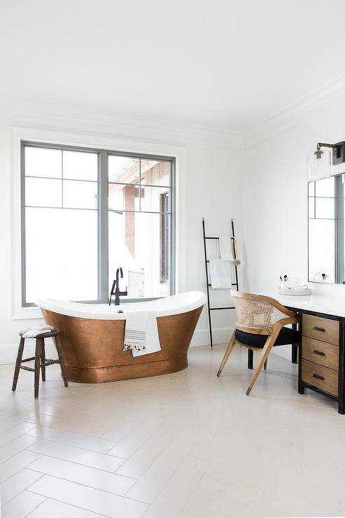 Copper bathtub fitted with a matte black tub filler under a window on an off white herringbone tiled floor in a transitional bathroom. A black towel ladder adds a stylish Scandinavian touch along wood and black cushion chair at a makeup vanity.