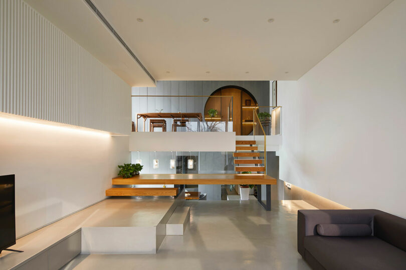 elongated living room with white walls and minimalist furnishings with a unique staircase at the far wall leading to mezzanine level