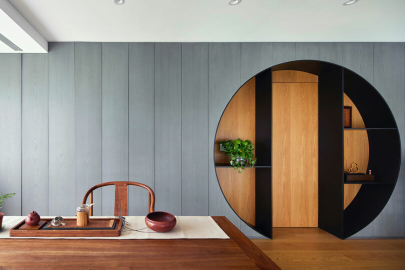 interior shot of partial dining table and chair with gray wall behind featuring a circular cutout with light wood door and shelving