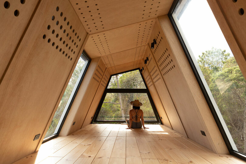 interior of minimalist cabin with wood walls with woman sitting facing away looking at trees