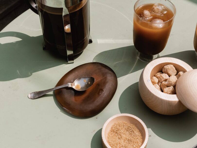 Material Kitchen Resting Stone in Cocoa holding a spoon, surrounded by coffee in a cup, sugar, and a coffee press