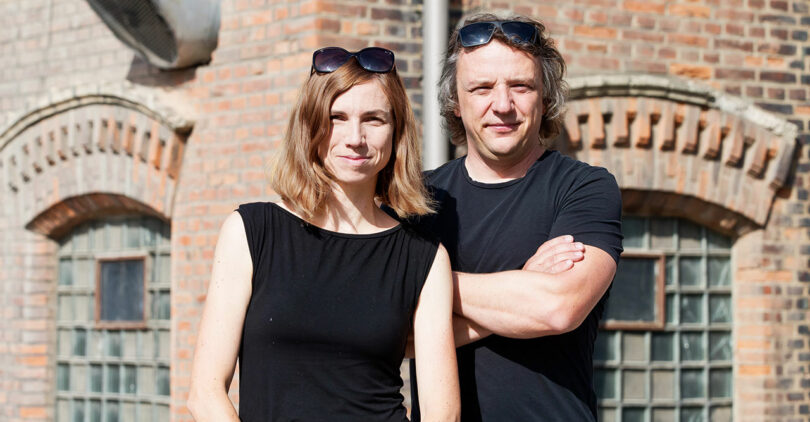 woman and man standing side by side in black shirts outside in front of a brick building