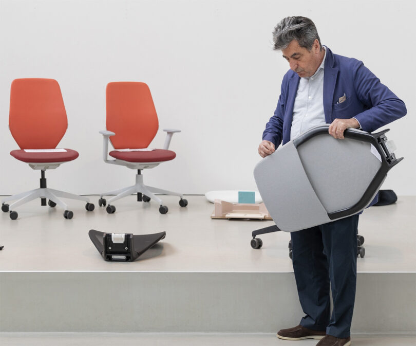 Designer Antonio Citterio in a blue jacket and slacks holding up the upper half of his Vitra ACX task chair with stretch fabric back. In the background are the other pieces of the chair, including its caster wheel base and seat, with two other ACX task chairs on the left in orange, red fabrics with light gray base with wheels.