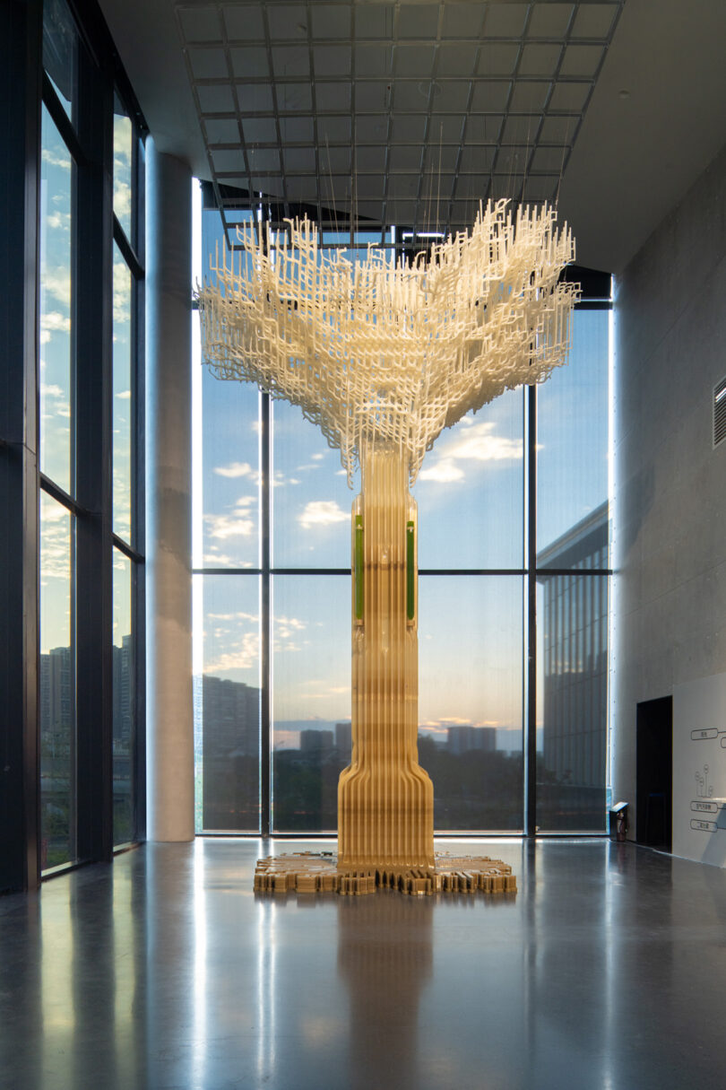 3D printed Tree.One carbon sequestering sculpture from the front. Shaped like a tree, the sculpture is shown displayed at the Chengdu Biennale 2023 inside a high ceiling exhibition space with a cityscape in the background seen through the buildings large scale windows.
