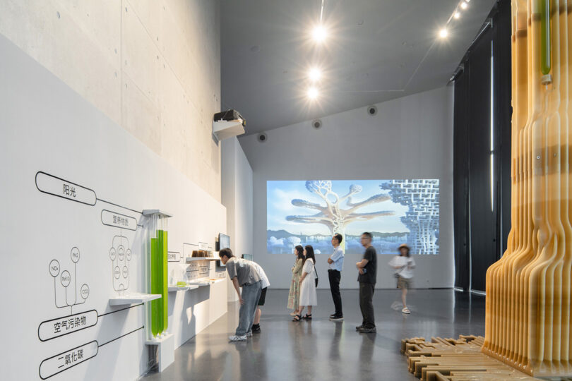 Small crowd of exhibition attendees near display stationed near Tree.One sculpture explaining the carbon sequestering process using algae in three clear tubes. Explanation is displayed in Chinese. A movie is projected with 3D animated city of future is projected onto the far wall.