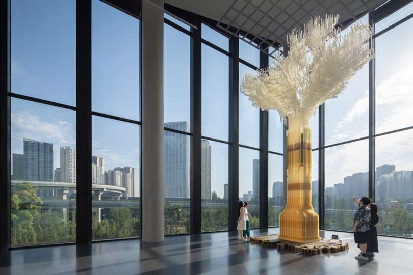 3D printed Tree.One carbon sequestering sculpture shaped like a tree displayed at the Chengdu Biennale 2023 inside a high ceiling exhibition space with a cityscape in the background seen through the buildings large scale windows.