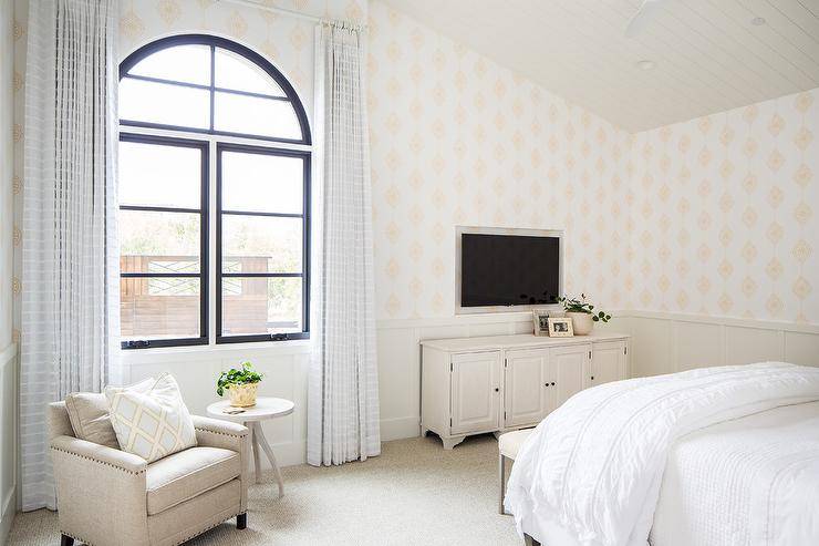 Restful guest bedroom features a television niche framed by white and gold wallpaper and positioned beneath a vaulted ceiling and over a vintage TV cabinet placed in front of light taupe wainscoting. Black framed arched windows are covered in long white striped curtains hung behind a beige accent chair placed beside a round rustic table.