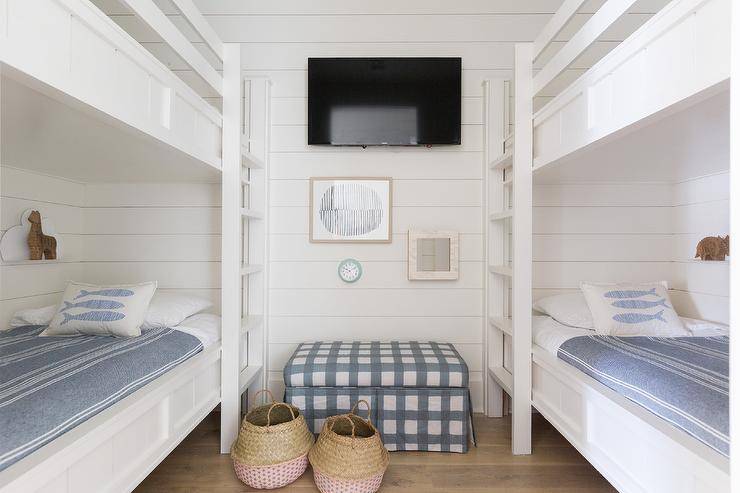 White and blue cottage bunk room boast white shiplap bunk beds dressed in white and blue bedding and fixed on opposite walls against a white shiplap trim. Between the beds, a blue and white gingham bench is positioned beneath a flat panel television hung over framed art.