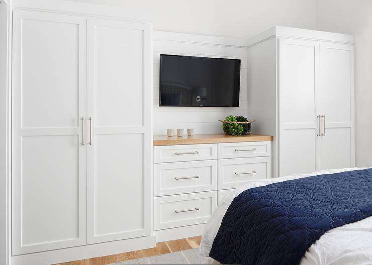 White built-in wardrobes flank a built-in dresser accented with a wood top and fixed beneath at flat panel television. The television is mounted facing a bed draped with a blue blanket.