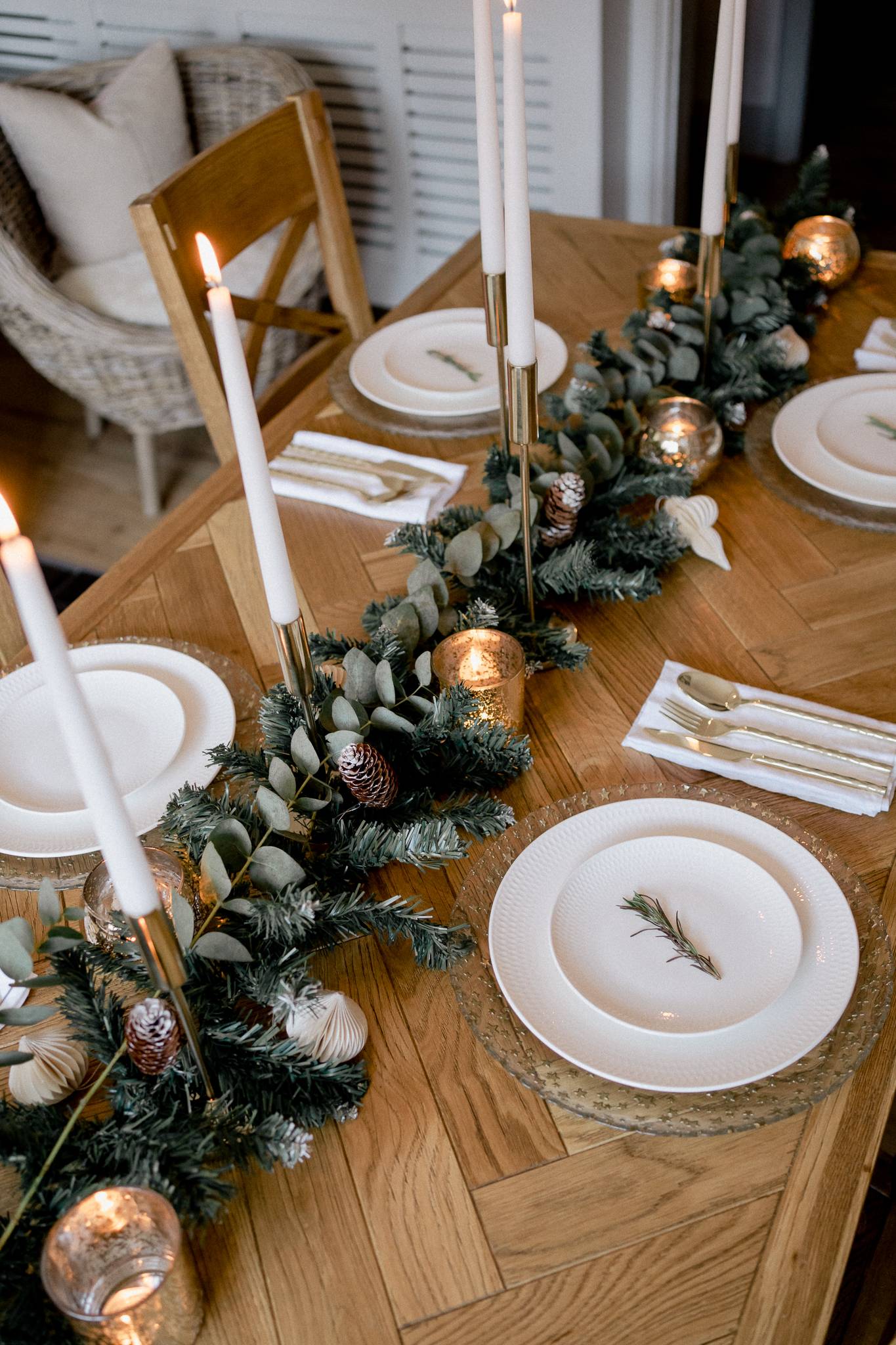 a glam winter tablescape with gold placemats, an evergreen, white bloom and snowy pinecone runner, candles and gold cutlery