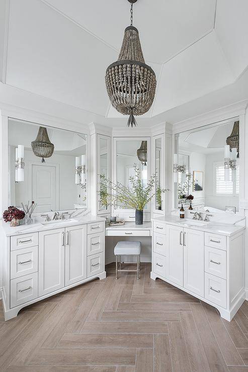 Hung from a vaulted ceiling over wood-like floor tiles, a gray beaded chandelier lights a well-appointed bathroom. A chrome vanity stool sits beneath a drop-down makeup vanity fitted in a corner and topped with a white quartz countertop mounted beneath white framed vanity mirrors. The makeup vanity is located between white shaker washstands boasting chrome pulls and white quartz countertops finished with chrome faucets mounted under white frame vanity mirrors.