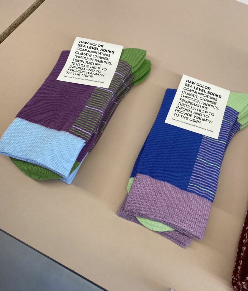 Two pairs of neatly folded socks – one is purple with a pale blue cuff and green toe and heel, the other is blue with a pale purple cuff and light green toe and heel. Both feature a series of horizontal lines up the ankle part and a cardboard label that reads "Raw Color Sea Level Socks communicating climate change through fabrics. Temperature textiles help to inform and provide warmth to the user.