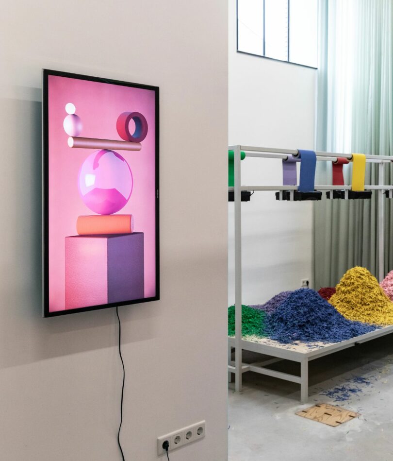 A vertically mounted TV screen shows a pink background with a delicately balanced pile of objects in different shades of pink, purple and orange. The paper shredding installation previous described can be seen to the right in the background. 
