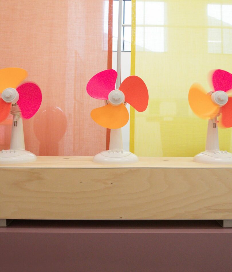 A series of three electric fans sit on a wooden box on a mauve plinth in front of an orange and a yellow curtain. Each fan has three blades – one pink, one dark orange and one yellowy-orange.