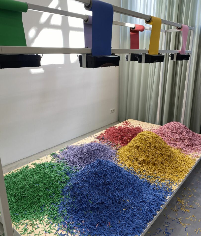 A series of paper shredder tops are attached to a metal frame with green, purple, blue, red, yellow and pink paper rolls going into them. Beneath confetti-like paper in the corresponding colors piles up on a low-level plinth.
