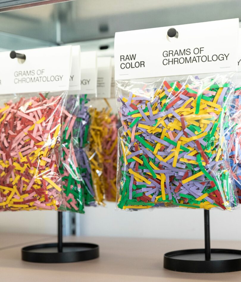 Small plastic bags of the shredded paper from the previous emails are sealed with a cardboard top reading "Raw Color Grams of Chromatology" and hung from retail display units in black metal. 