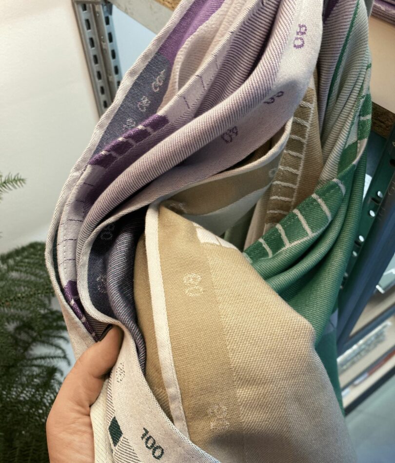 A white women's hand hold a handful of tea towels showing the different colours (purple, beige and green) and the reference codes down the edge. 