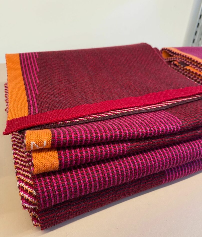 A pile of neatly folded purple, red, orange and pink scarves feature lines of differing lengths.
