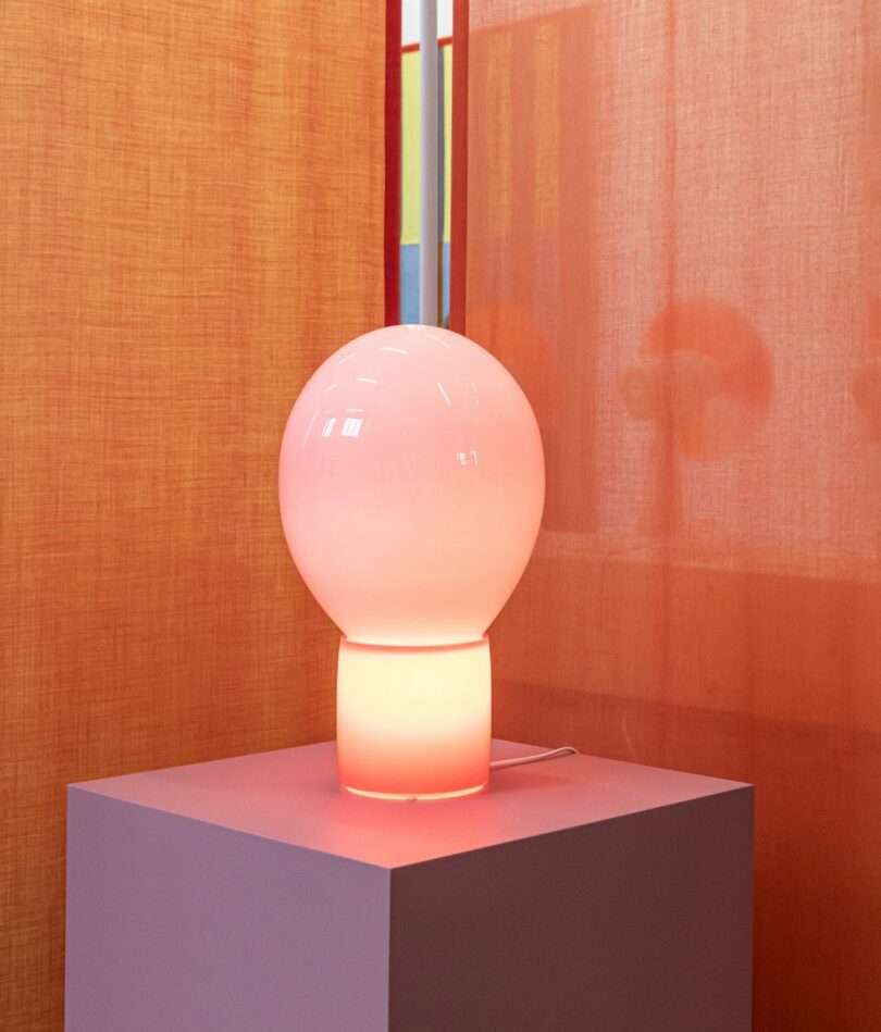 A pink and orange glass lamp is formed on a cylindrical base and a balloon-shaped shade. It stands on a mauve plinth in front of an orange curtain. 