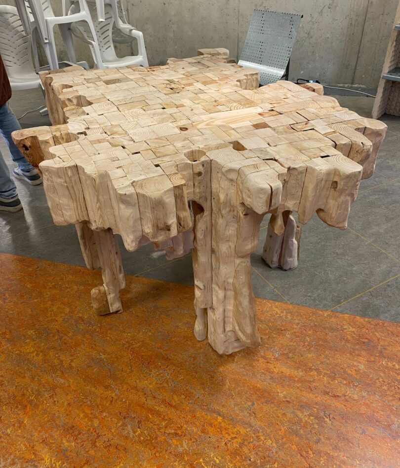 A sculptural table is made from vertical segments of wood. It has a very uneven, wavy edge, a very deep surface and multiple, irregular legs.