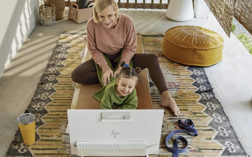 Mother with daughter using HP Envy Move computer from the ground. Mother is seated with daughter laying on her stomach on the ground using/viewing the computer. The pair are in a room that appears both indoors and partially outdoors set up for yoga, with floor pillows, rug and yoga mat.