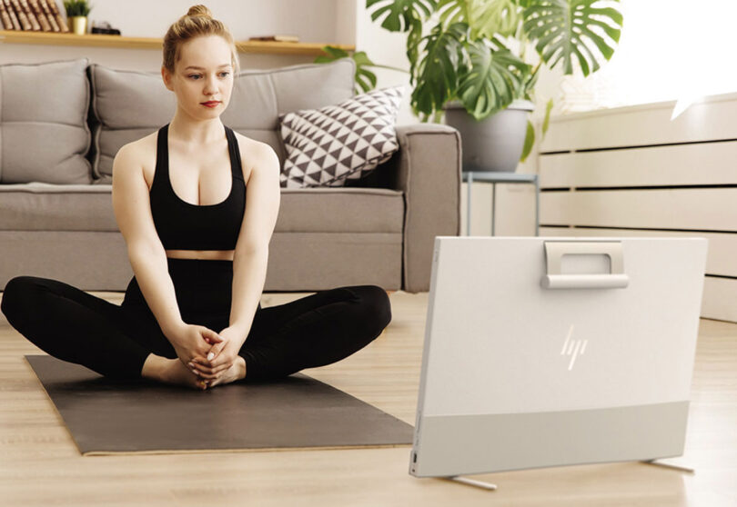 Young woman in black workout clothing on the ground in seated yoga pose on a yoga mat looking at HP Envy Move computer in a living room setting with sofa and houseplants in the background. Back of computer includes a handle and HP logo.