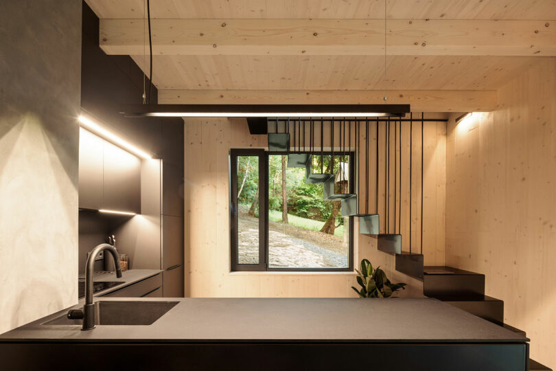 view into modern black kitchen with light wood ceiling and simple staircase behind