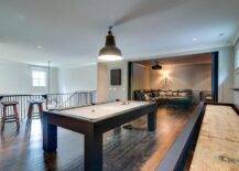 Ideal for entertainers, this game room is designed on a second floor in a large home in a darker rugged wood theme. Black industrial flush mount lights hang over a large landing pool table and movie room. Long shuffleboard is a highlight in this game room adding to the textural wood accents such as a dark stained plank wood floor. A gray curved leather sectional offers comfortable seating in a tv room while the game room displays industrial styled barstools.