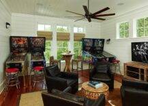 Dream Man Cave style games room with pinball machines and fifties style chrome bar stools flanking three windows. Below the windows stands a pub height wooden table paired with seagrass bar stools. In the foreground four espresso leather club chairs with nailhead trim surround a round wooden drum coffee table. The seating arrangement is anchored by a brown and white cowhide rug layered on top of a bound sisal rug over hardwood floors. To the right of the seating arrangement stands a distressed wood media console with flat screen tv. The room features white shiplap paneled walls and wood paneled ceilings. A contemporary ceiling fan adorns the ceiling over the space.